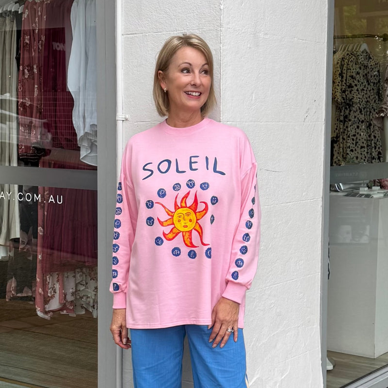 This pink sweater has a round neckline, drop shoulder, long sleeves with ribbed cuffs and 'Soleil' in navy on the front along with a sun and navy Zodiac signs surrounding it.