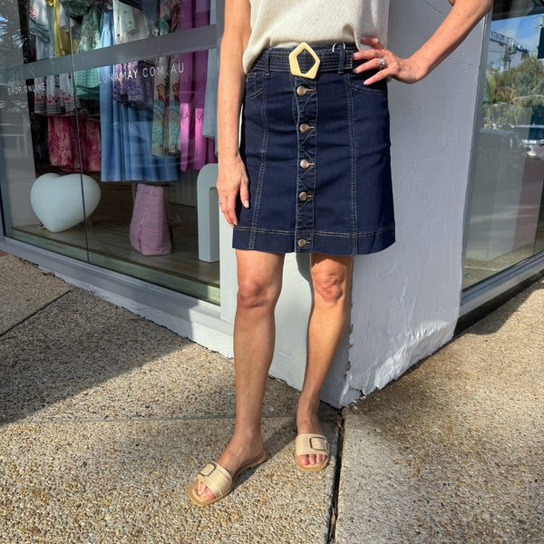 Our Theo Denim Skirt is available in a dark indigo wash. This mini skirt has a  gold button front and a detachable self fabric belt with a gold buckle. It also has front pockets.
