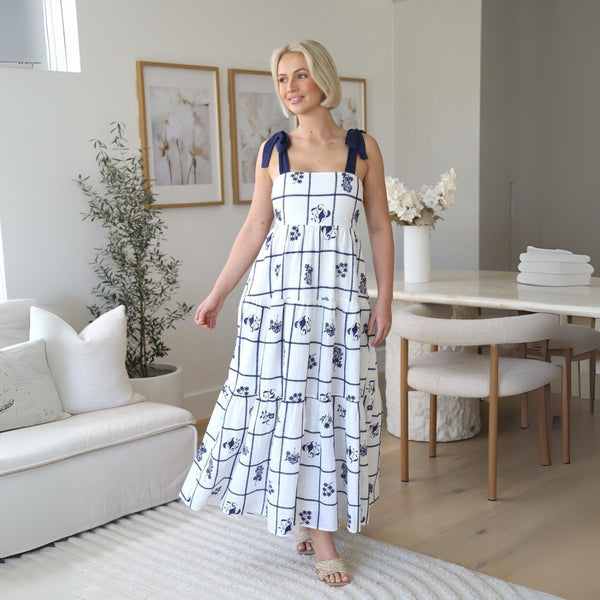 Our Britney Maxi Dress has a white base with an embroidered navy floral design and navy check stripes. 