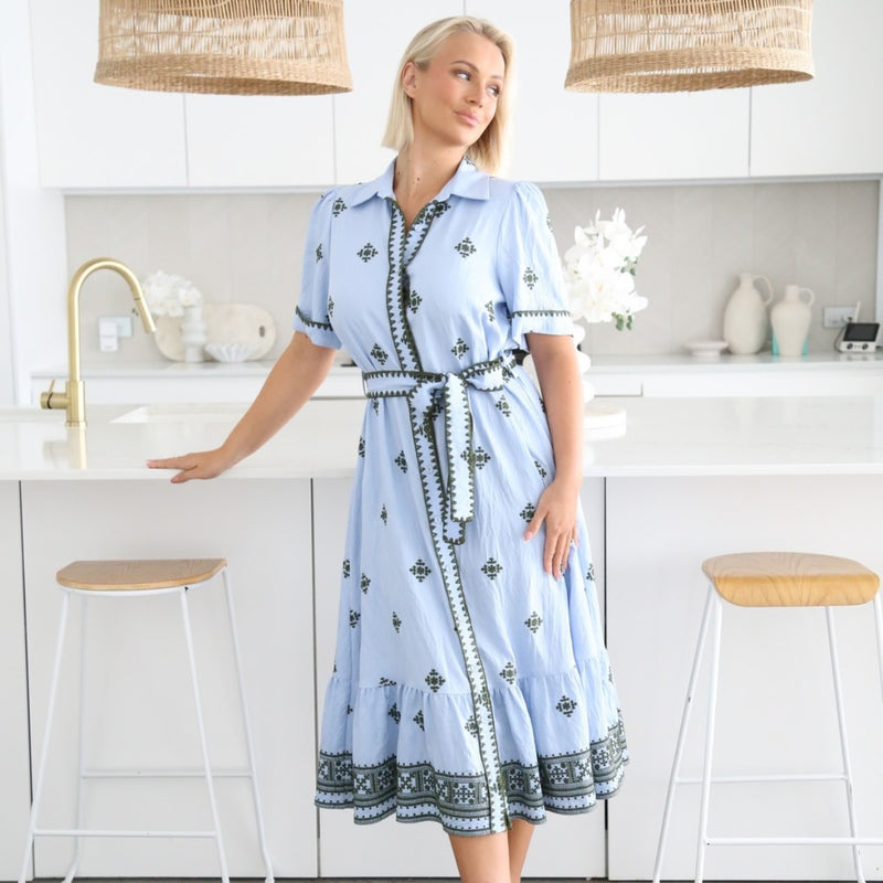 Our Delaney Shirt Dress has a powdery blue base with khaki embroidery detailing. The dress features a collar, short sleeves, button front and it has a frill hem.